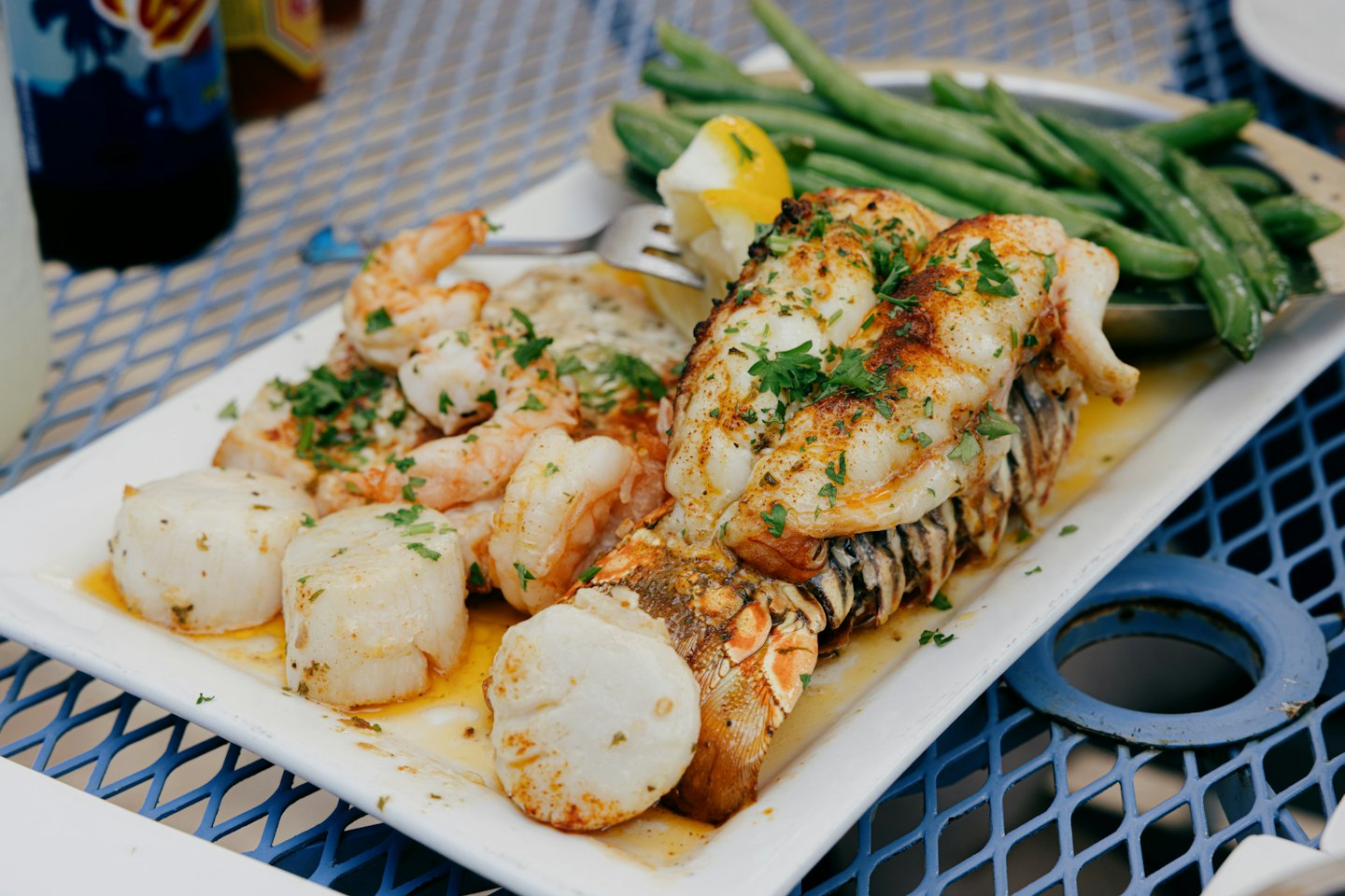 Seafood platter with lobster, shrimp, scallops, and green beans on a blue mesh table.
