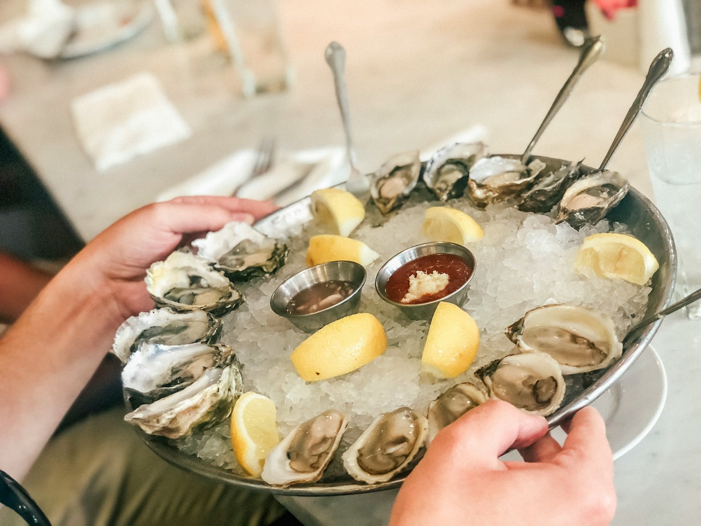 Hands holding a platter of shucked oysters with lemon and cocktail sauce.